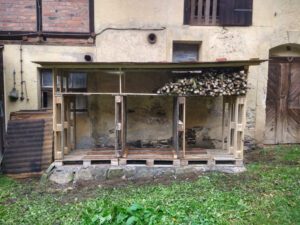 Eine Regalartige Konstruktion aus dem Projekt "bauen mit Paletten". Sie besitzt ein Dach, das mit Ziegelsteinen beschwert ist. Ein Fach ist schon teilweise mit Brennholz befüllt.