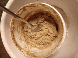 Ein rundes, weißes Gefäß mit Teig darin. In dem Teig steckt ein Esslöffel. Die Oberfläche des Teiges wirkt angetrocknet.