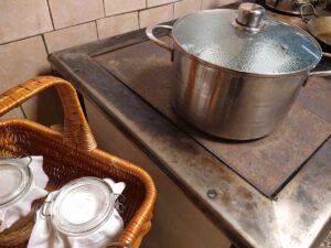Rechts ist ein Holzherd zusehen, auf dem ein großer Edelstahl Kochtopf mit Glasdeckel steht. Links steht ein Tragekorb mit Einkochgläsern, welche mit weißen Tüchern abgedeckt sind. Der Glasdeckel des Topfes ist mit Feuchtigkeit beschlagen, offenbar ist der Herd heiß. Gläser sterilisieren ist der Zweck des Aufbaus.